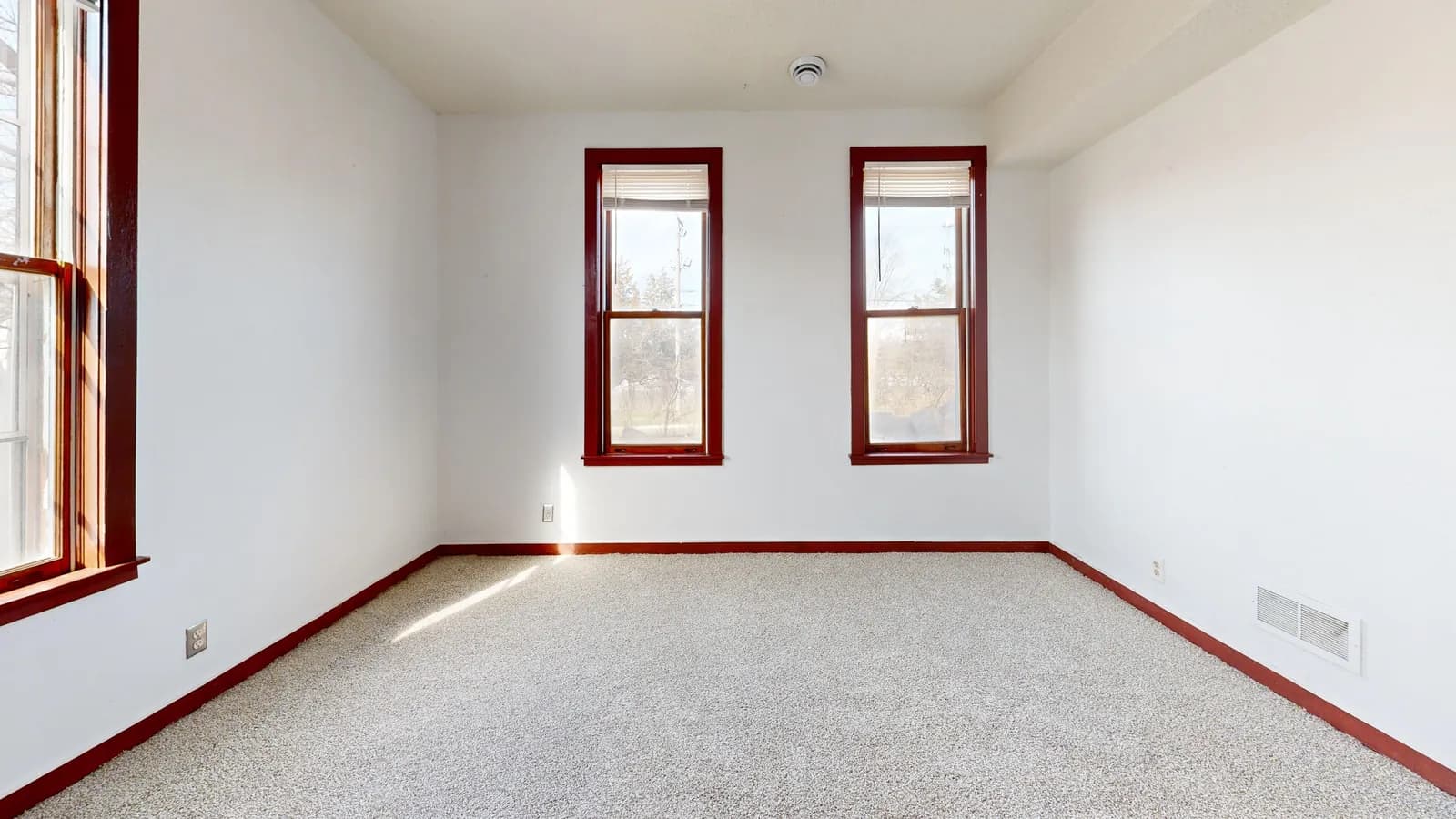 An empty carpeted room lit by sunlight through two windows