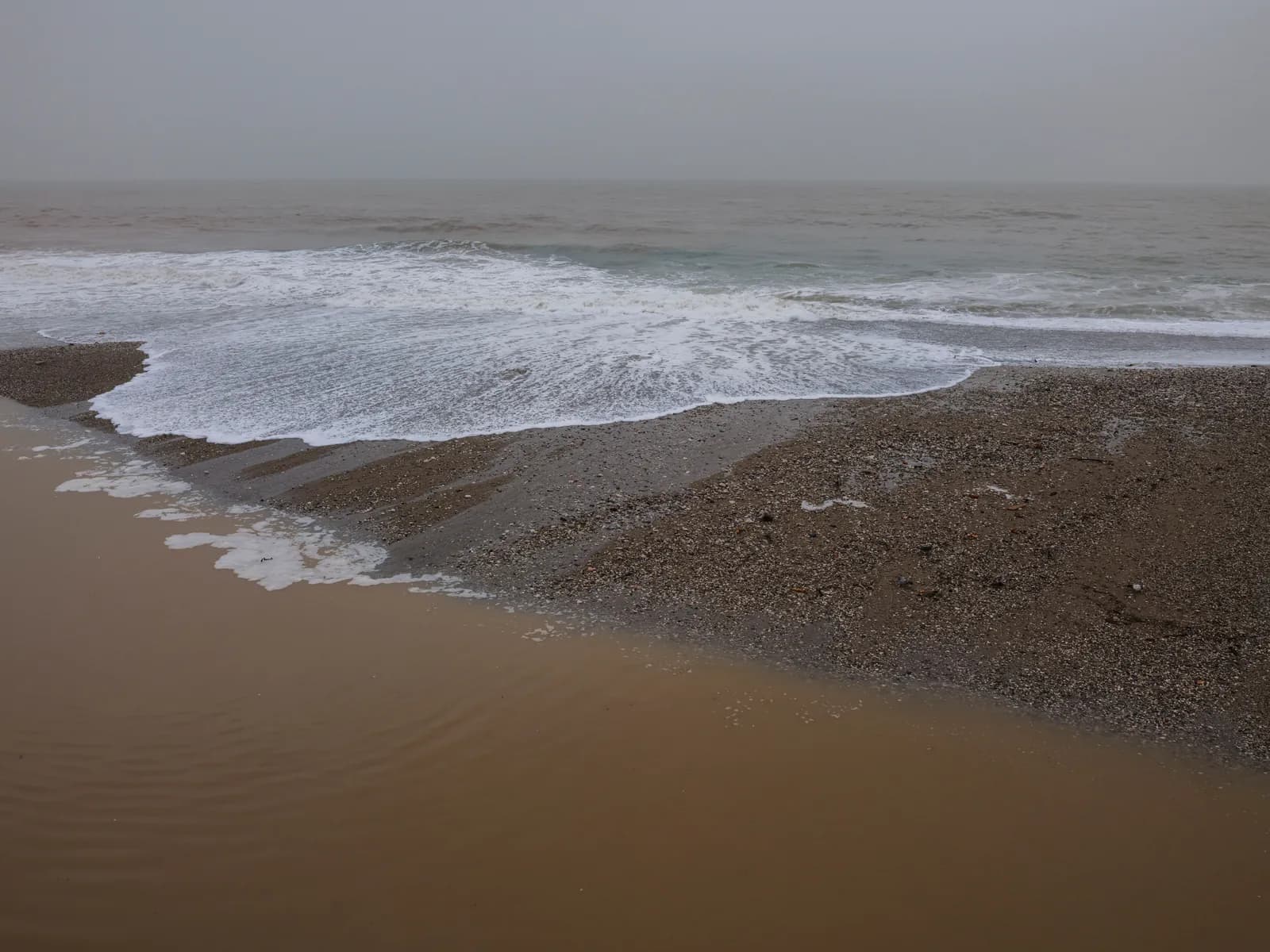 Grey surf pushing up a flat sand beach under an overcast sky