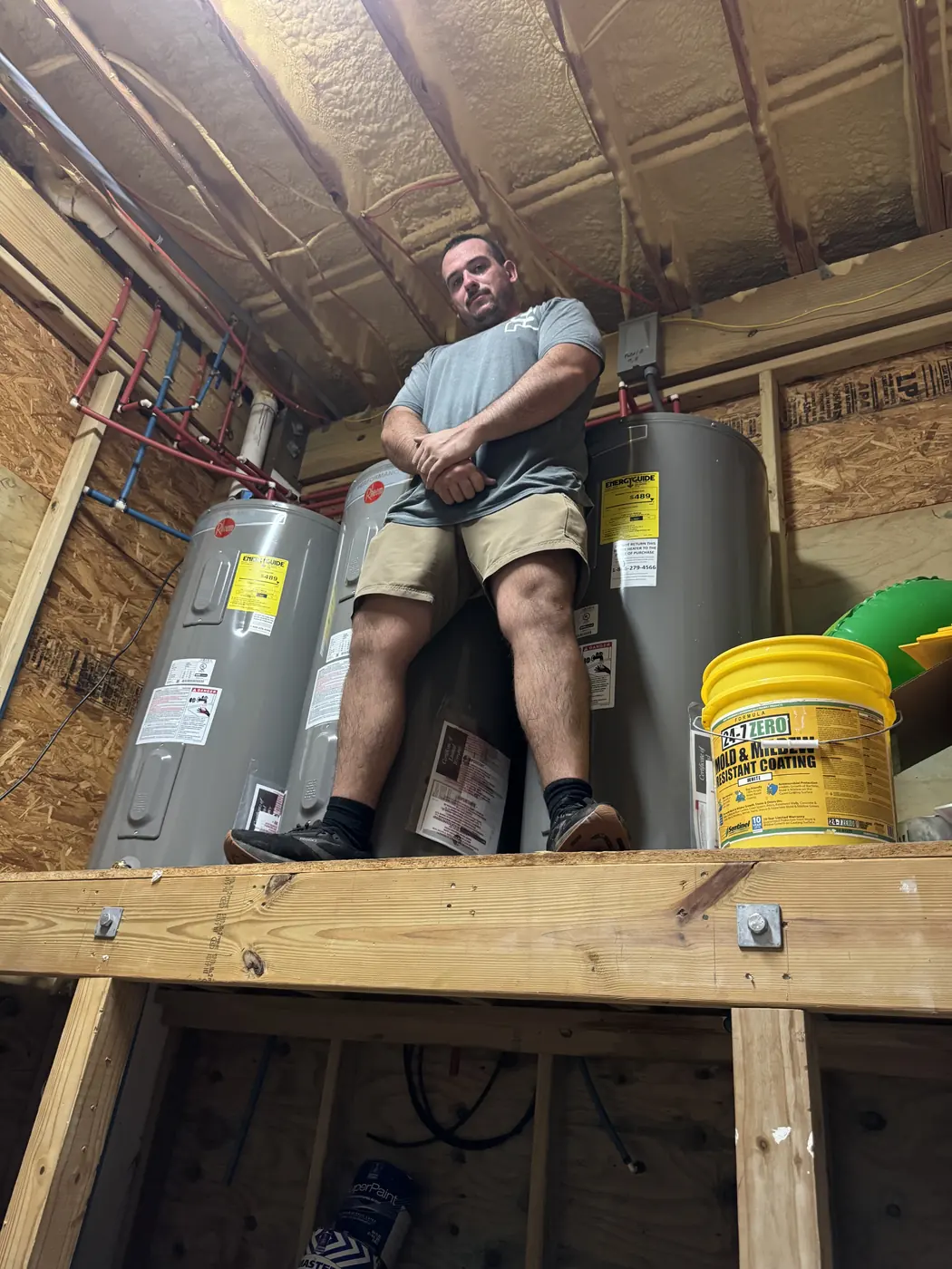 Streamlined Plumbing technician beside three tank water heaters in a framed mechanical room.